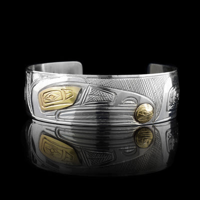 This 14K gold and sterling silver cuff bracelet has a wide band with a gap in the back. There are carvings on the surface of the band that depict the Raven holding a pearl. The eye of the raven and the pearl are made from gold and also have unique carvings on it.