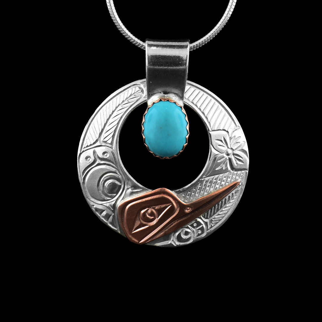This turquoise pendant has a sterling silver circle with a circle cutout in the center of it. The head of the Hummingbird is done in the bottom using copper and the details of the head are carved into it. On the left of the piece, the wing details are depicted and on the other side there is a flower.
There is an oval turquoise gem in the center of the cutout that is held in copper and there is a wide, silver bail above the gem.