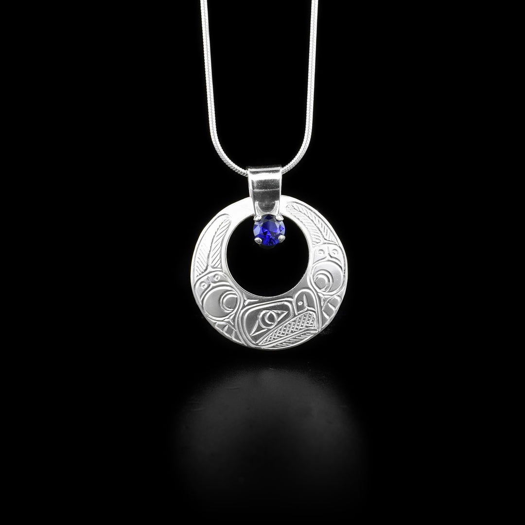 This sterling silver gemstone pendant is circle shaped and has a circle cutout in it with a lab grown blue gem at the top. There is a depiction of the Raven carved into the surface of the pendant.
