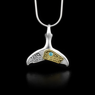 This Orca whale tail pendant is made form sterling silver, 14K gold and turquoise lab grown gemstone. The pendant is shaped like a whale tail and has carvings on the surface of it that depict the Orca. The head of the Orca is made form the gold and the gem has been placed in its eye.