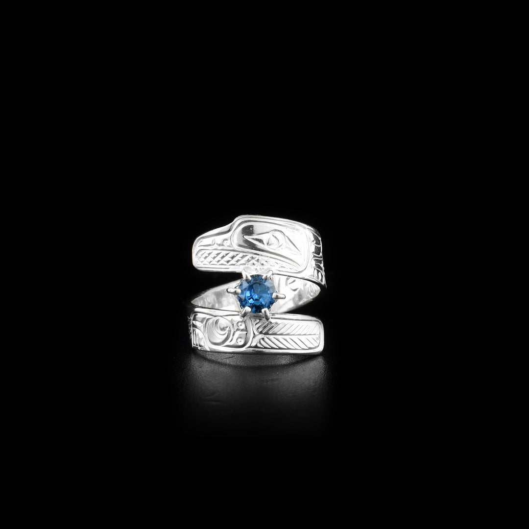 This blue gemstone ring is made from sterling silver and has a wrapped, tapered band. There is a depiction of a flower and the Raven carved into the surface of it.