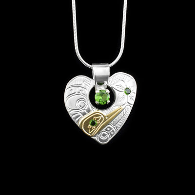 This silver and gold heart pendant is made from sterling silver, 18K gold and green lab grown gemstone. The pendant is heart shaped and has a small cutout with a green gemstone in it. There are carvings on the surface of the pendant that depict the Hummingbird and its head is made from 14K gold.