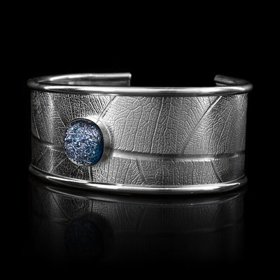 This beautiful leaf pattern, wide cuff bracelet is made using sterling silver and a denim (blue and white speckled) colored fused glass bead. The piece has a single, wide band with a gap in the back and there are detailed leaf textures imprinted across the surface of it. The glass bead has interesting details on the surface of it and is embossed in the top, center of the bracelet
