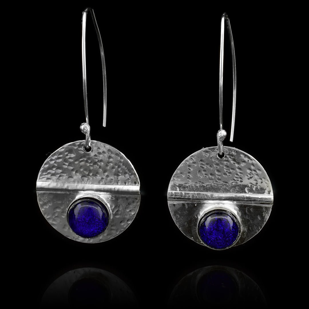 These dangling circle drop earrings are made from sterling silver and royal blue colored fused glass beads. The silver hangs are circle shaped and dangle from long, curved hoop hooks. The glass beads are held below a horizontal crease running through the hangs. There are dimpled textures made across the surface of the hangs.