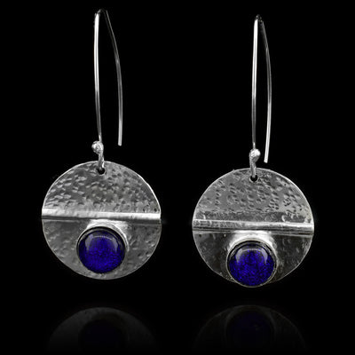 These dangling circle drop earrings are made from sterling silver and royal blue colored fused glass beads. The silver hangs are circle shaped and dangle from long, curved hoop hooks. The glass beads are held below a horizontal crease running through the hangs. There are dimpled textures made across the surface of the hangs.