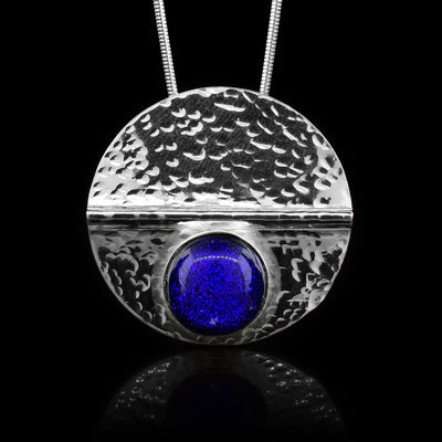 This sterling silver circle pendant has a dark blue colored fused glass bead embossed in the bottom half of the piece. There is a straight crease made horizontally through the piece and there are dimpled textures made all across the piece.