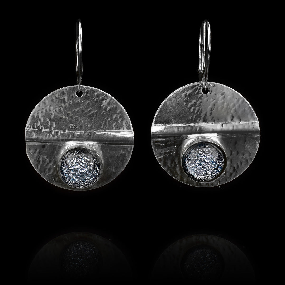 These circle dangle earrings have been made using sterling silver and denim patterned fused glass beads. The hangs are circle shaped and have dimpled textures on them and a crease running through center of them. The denim patterned beads are embossed into the bottom of the hangs.