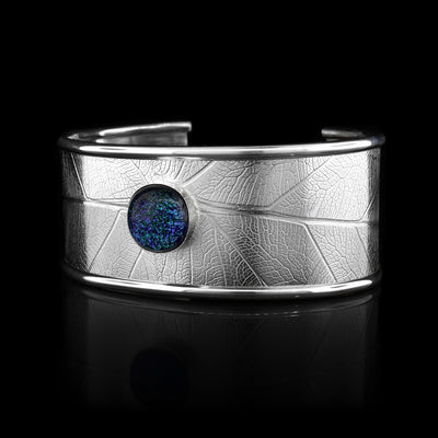 This beautiful leaf cuff bracelet is made using sterling silver and a turquoise colored fused glass bead. The piece has a single, wide band with a gap in the back and there are detailed leaf textures imprinted across the surface of it. The glass bead has interesting details on the surface of it and is embossed in the top, center of the bracelet