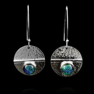 These dangling circle drop earrings are made from sterling silver and green colored fused glass beads. The silver hangs are circle shaped and dangle from long, curved hoop hooks. The glass beads are held below a horizontal crease running through the hangs. There are dimpled textures made across the surface of the hangs.