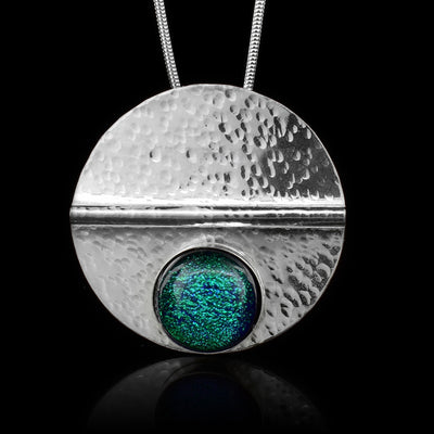 This sterling silver circle pendant has a green colored fused glass bead embossed in the bottom half of the piece. There is a straight crease made horizontally through the piece and there are dimpled textures made all across the piece.