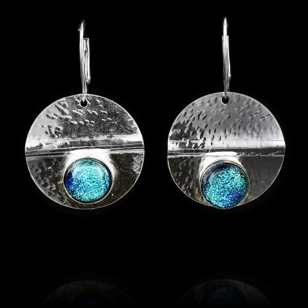 These circle dangle earrings have been made using sterling silver and light blue colored fused glass beads. The hangs are circle shaped and have dimpled textures on them and a crease running through center of them. The light blue beads are embossed into the bottom of the hangs.