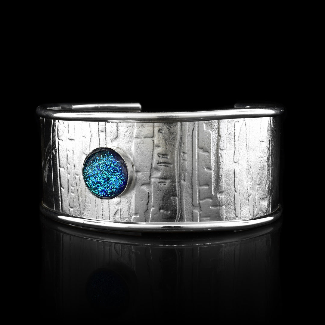 This wide cuff bracelet has has a single, wide band made from sterling silver and has a gap in the back. The edges of the band are raised and there are striped and abstract designs that have been pressed into the surface of it. There is a light blue colored fused glass bead that is embossed into the top of the band.