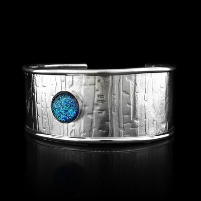 This wide cuff bracelet has has a single, wide band made from sterling silver and has a gap in the back. The edges of the band are raised and there are striped and abstract designs that have been pressed into the surface of it. There is a light blue colored fused glass bead that is embossed into the top of the band.