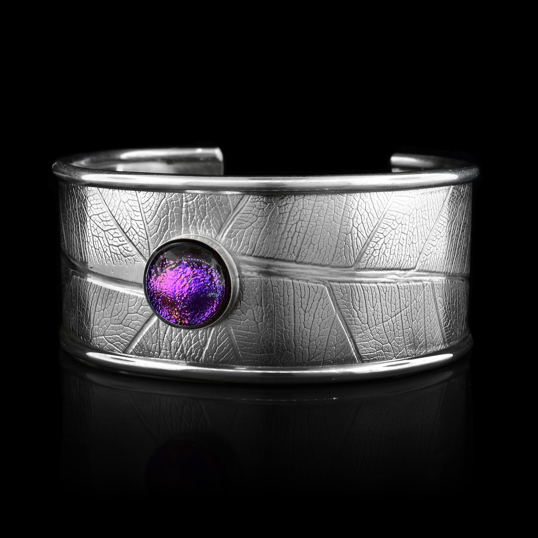 This beautiful leaf cuff bracelet is made using sterling silver and a purple colored fused glass bead. The piece has a single, wide band with a gap in the back and there are detailed leaf textures imprinted across the surface of it. The glass bead has interesting details on the surface of it and is embossed in the top, center of the bracelet