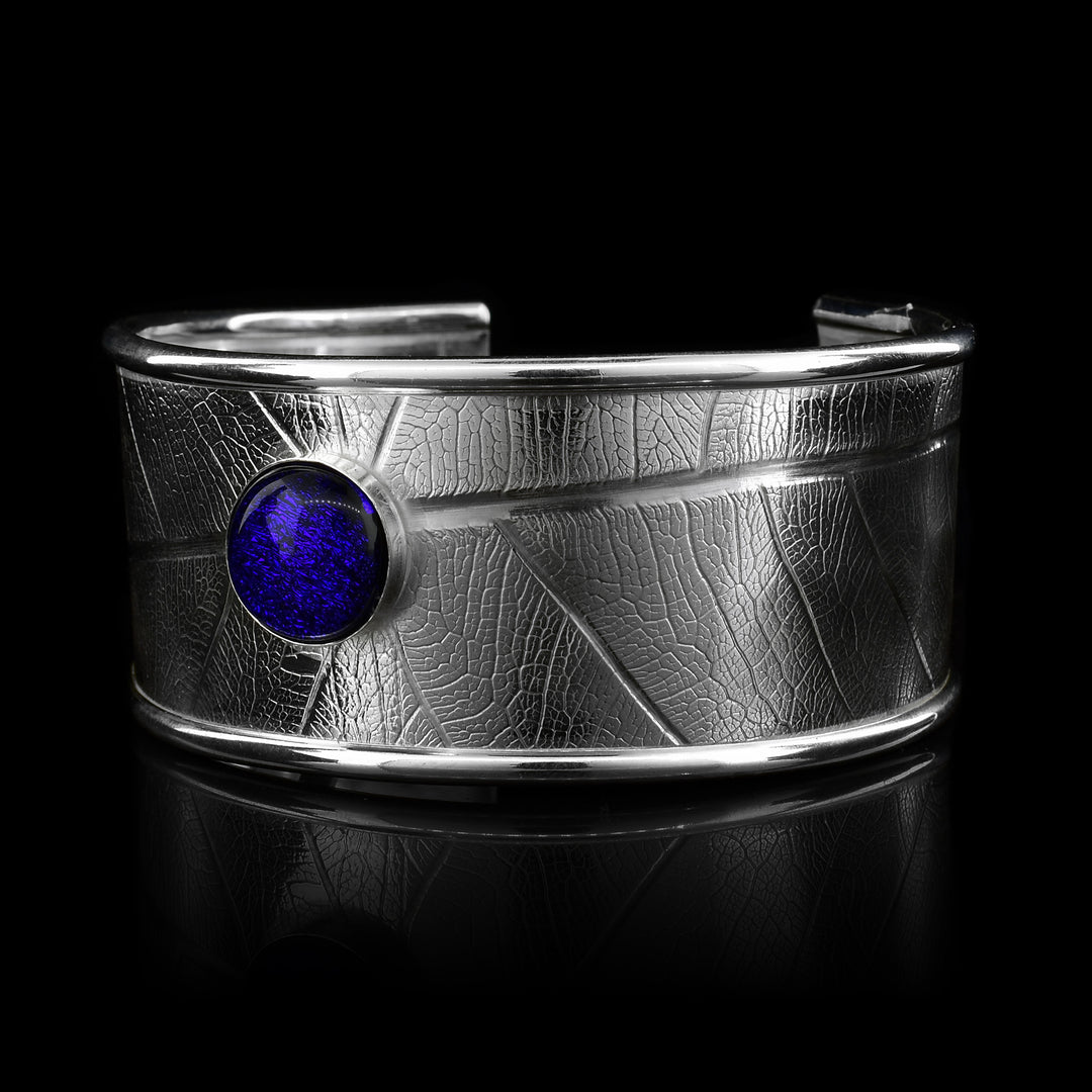 This beautiful leaf pattern, wide cuff bracelet is made using sterling silver and a royal blue colored fused glass bead. The piece has a single, wide band with a gap in the back and there are detailed leaf textures imprinted across the surface of it. The glass bead has interesting details on the surface of it and is embossed in the top, center of the bracelet