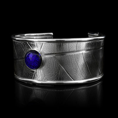 This beautiful leaf pattern, wide cuff bracelet is made using sterling silver and a royal blue colored fused glass bead. The piece has a single, wide band with a gap in the back and there are detailed leaf textures imprinted across the surface of it. The glass bead has interesting details on the surface of it and is embossed in the top, center of the bracelet