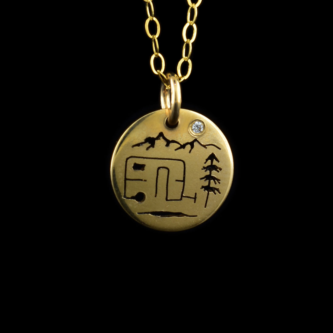 This camper and mountain pendant necklace has a small, circle pendant dangling from a jump ring on a simple chain. These pieces are 14K gold filled material and there is a silhouette of a camping scene on the pendant. In the middle there is a rectangle camper van and a tree right beside it. At the top of the pendant are two mountain peaks and a cubic zirconia gem at the moon in the sky.