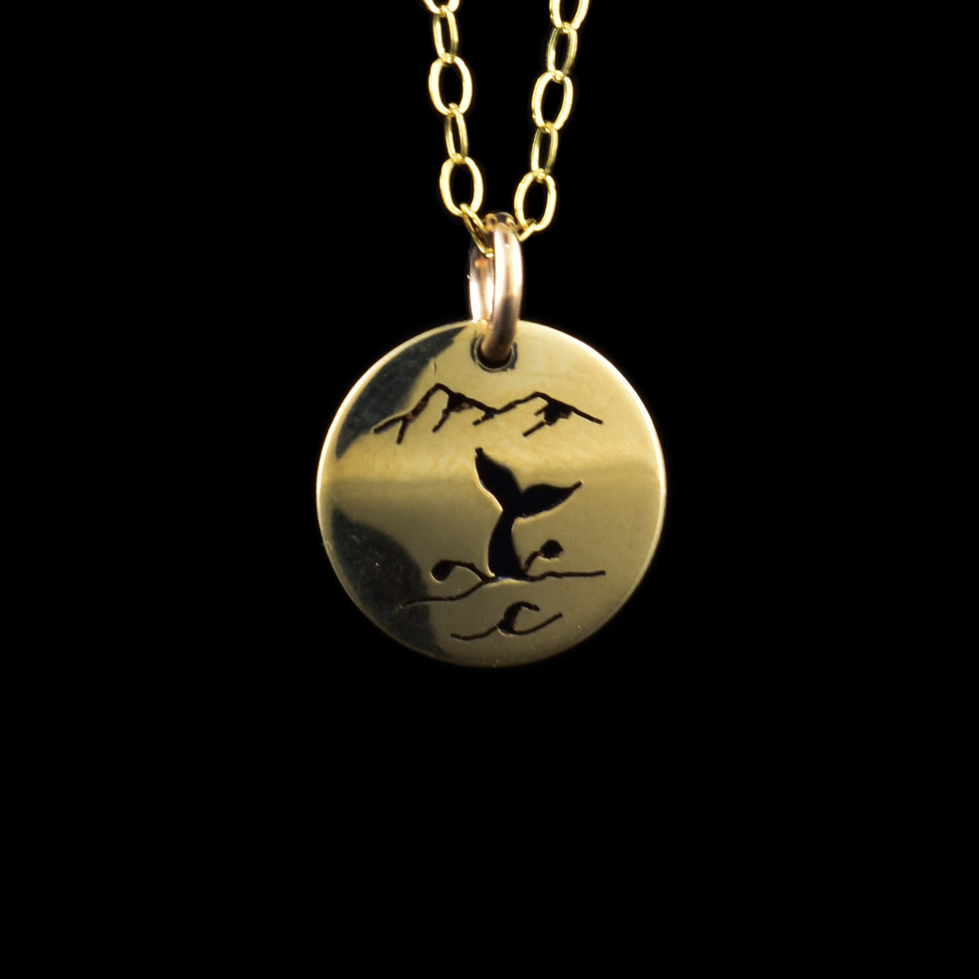 This whale tail pendant necklace has a small, circle pendant dangling from a jump ring on a simple chain. These pieces are made from 14K gold filled material and there is a ocean scene on the pendant. There are silhouettes are two mountain peaks on top of the piece and there is a whale tail coming out from the waves with splashing designs coming out of the side.
