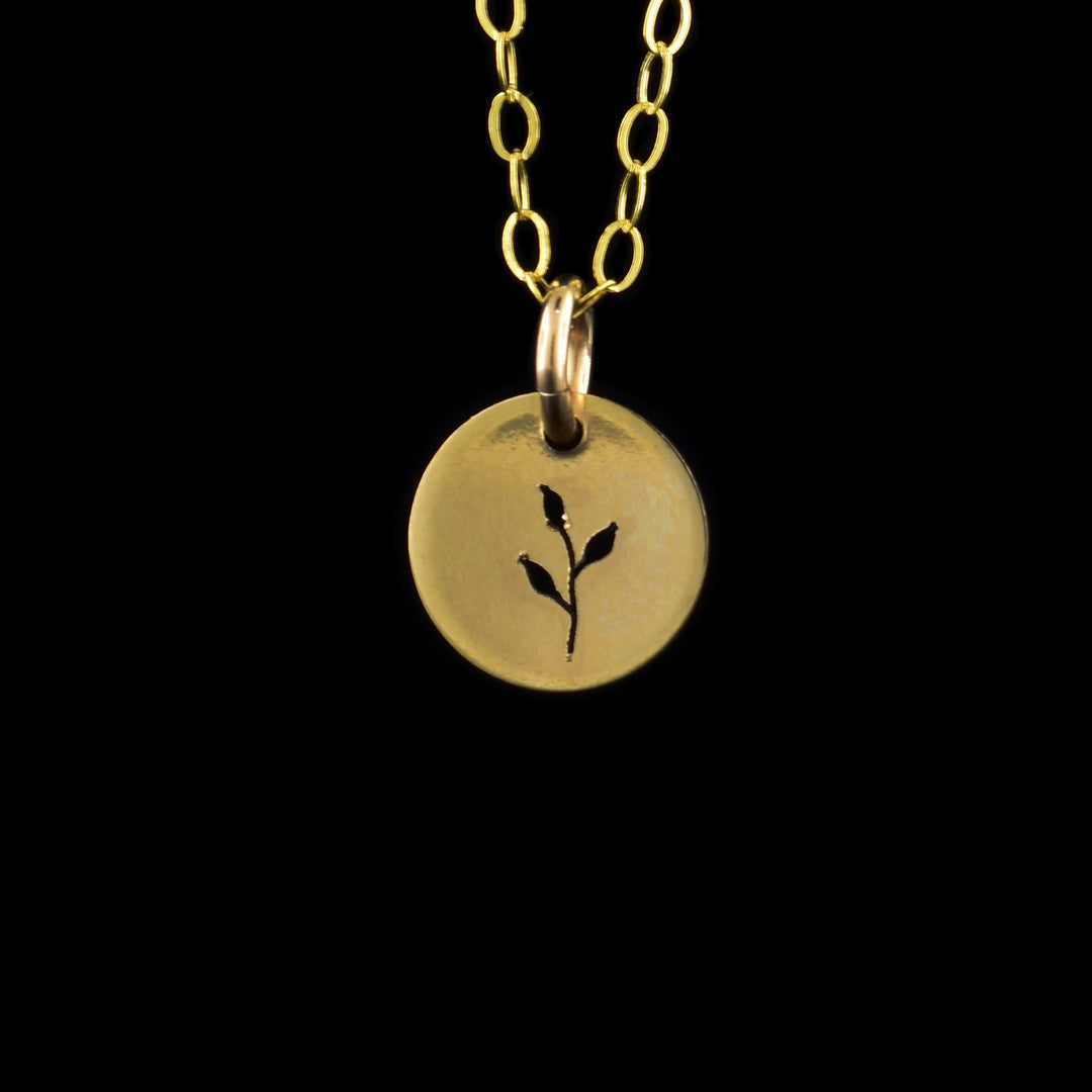 This leaf necklace pendant has a small, circle pendant dangling from a jump ring and a simple chain. These pieces are made from 14K gold filled material and there is a cutout design on the pendant. There is a small sprout with a stem that curves slightly and has three leaves extending from it.