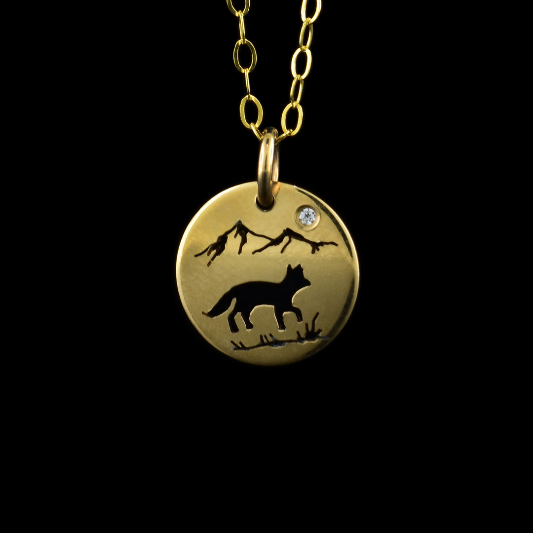 This Fox pendant necklace has a small, circle pendant dangling from a jump ring on a simple chain. These pieces are made from 14K gold filled material and there is a silhouette scene of a fox in a mountain terrain. There are two peaks with a cubic zirconia gem above them with a side view of a fox below. There is a line with grass under the fox.