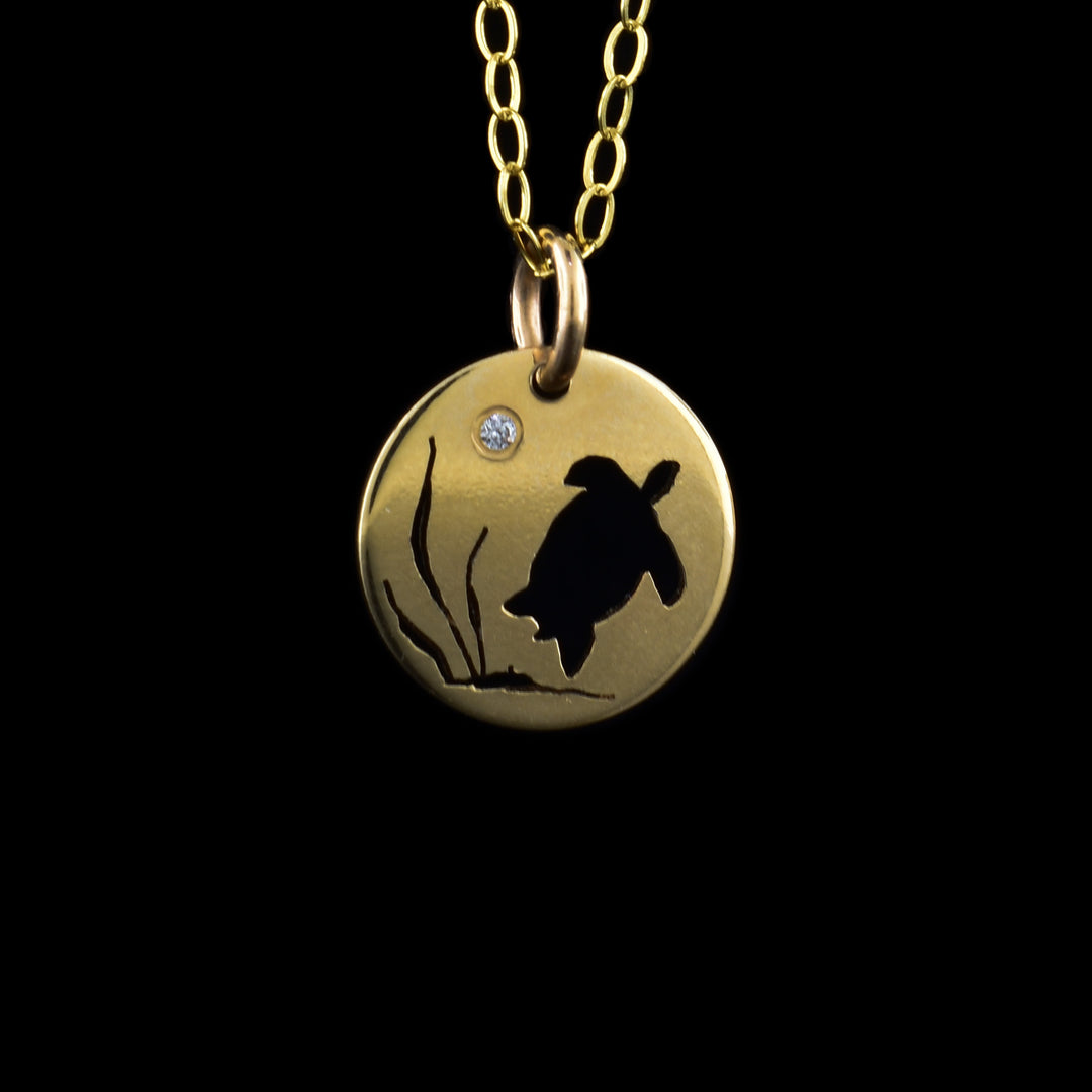 Sea Turtle Pendant Necklace has a small, circle pendant dangling from a jump ring on a simple chain. These pieces are made from 14K gold filled material and there is a silhouette of a sea turtle in kelp. On the left there are three strands of kelp and there is a large sea turtle with its shell facing the viewer. There is a cubic zirconia gem at the top of the pendant representing the moon.
