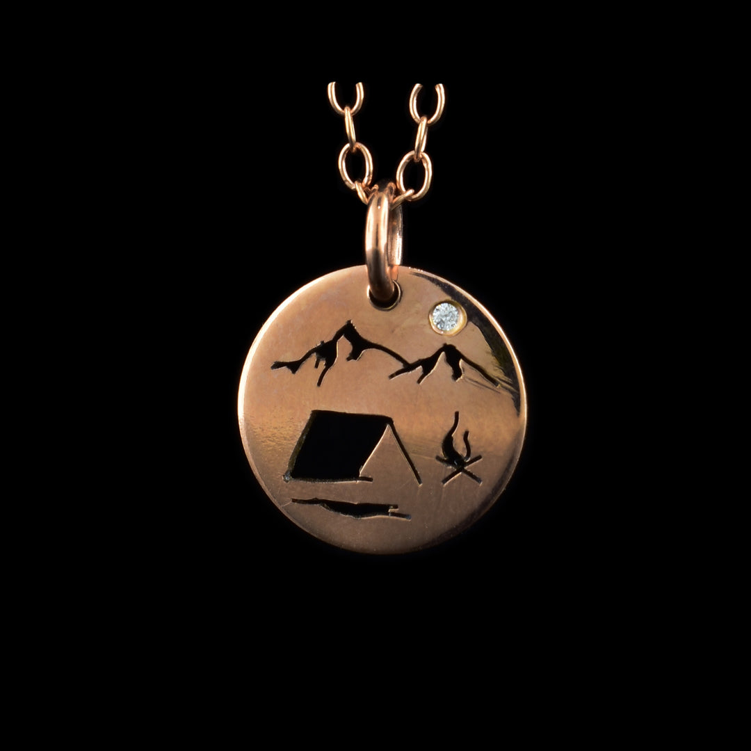 This rose gold necklace and pendant has a small, circle pendant dangling from a jump ring on a simple chain. These pieces are made from 14K rose gold filled material and there is a silhouette scene of a campsite. There is a simple tent at the bottom with a campfire to the right of it. At the top of the piece, there are two mountain peaks with a cubic zirconia gemstone in the "Sky".