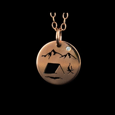 This rose gold necklace and pendant has a small, circle pendant dangling from a jump ring on a simple chain. These pieces are made from 14K rose gold filled material and there is a silhouette scene of a campsite. There is a simple tent at the bottom with a campfire to the right of it. At the top of the piece, there are two mountain peaks with a cubic zirconia gemstone in the "Sky".