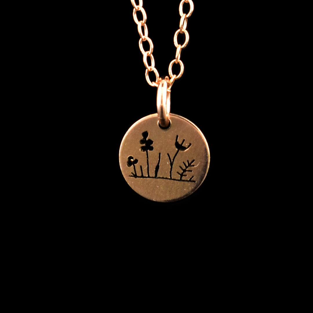 This rose gold necklace and pendant has a small, circle pendant dangling form a jump ring on a simple chain. These pieces are made from 14K rose gold filled material and there are wild flower designs made on the pendant. From left to right, there is a small clover, a larger lilac, a daisy and another smaller fern.