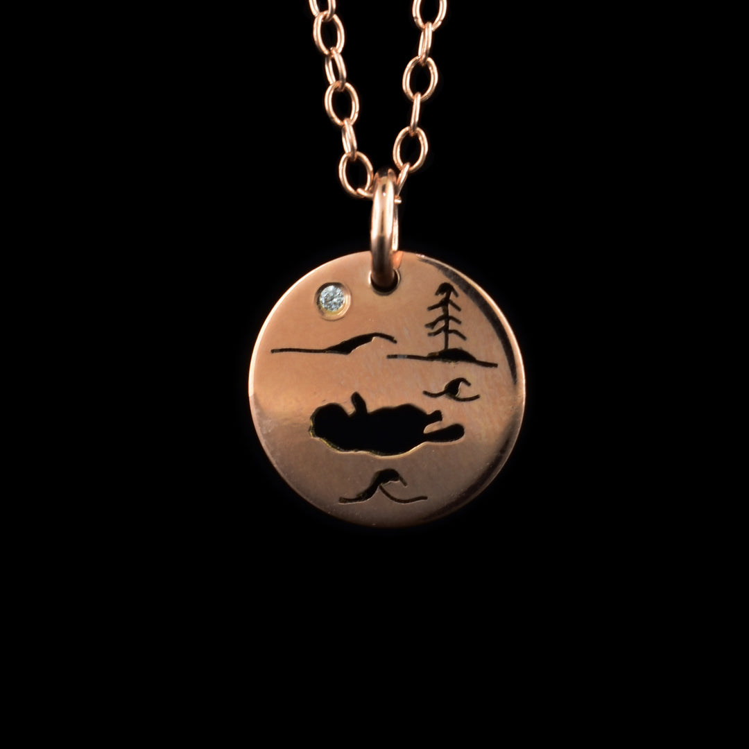 This rose gold necklace and pendant has a small, circle pendant dangling from a jump ring on a simple chain. These pieces are made from 14K rose gold and there is a depiction of a sea otter in its natural habitat. The scene is a silhouette and shows the otter on its back with wave designs around it. There is a mountain and a tree in the background a cubic zirconia representing the moon.