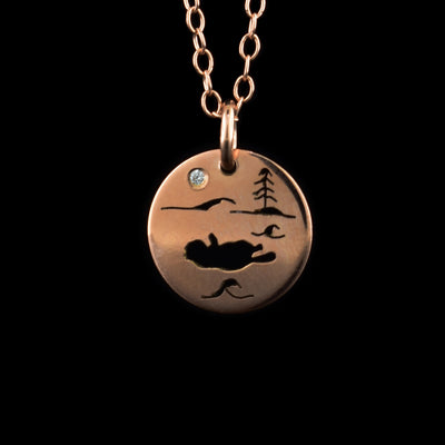 This rose gold necklace and pendant has a small, circle pendant dangling from a jump ring on a simple chain. These pieces are made from 14K rose gold and there is a depiction of a sea otter in its natural habitat. The scene is a silhouette and shows the otter on its back with wave designs around it. There is a mountain and a tree in the background a cubic zirconia representing the moon.
