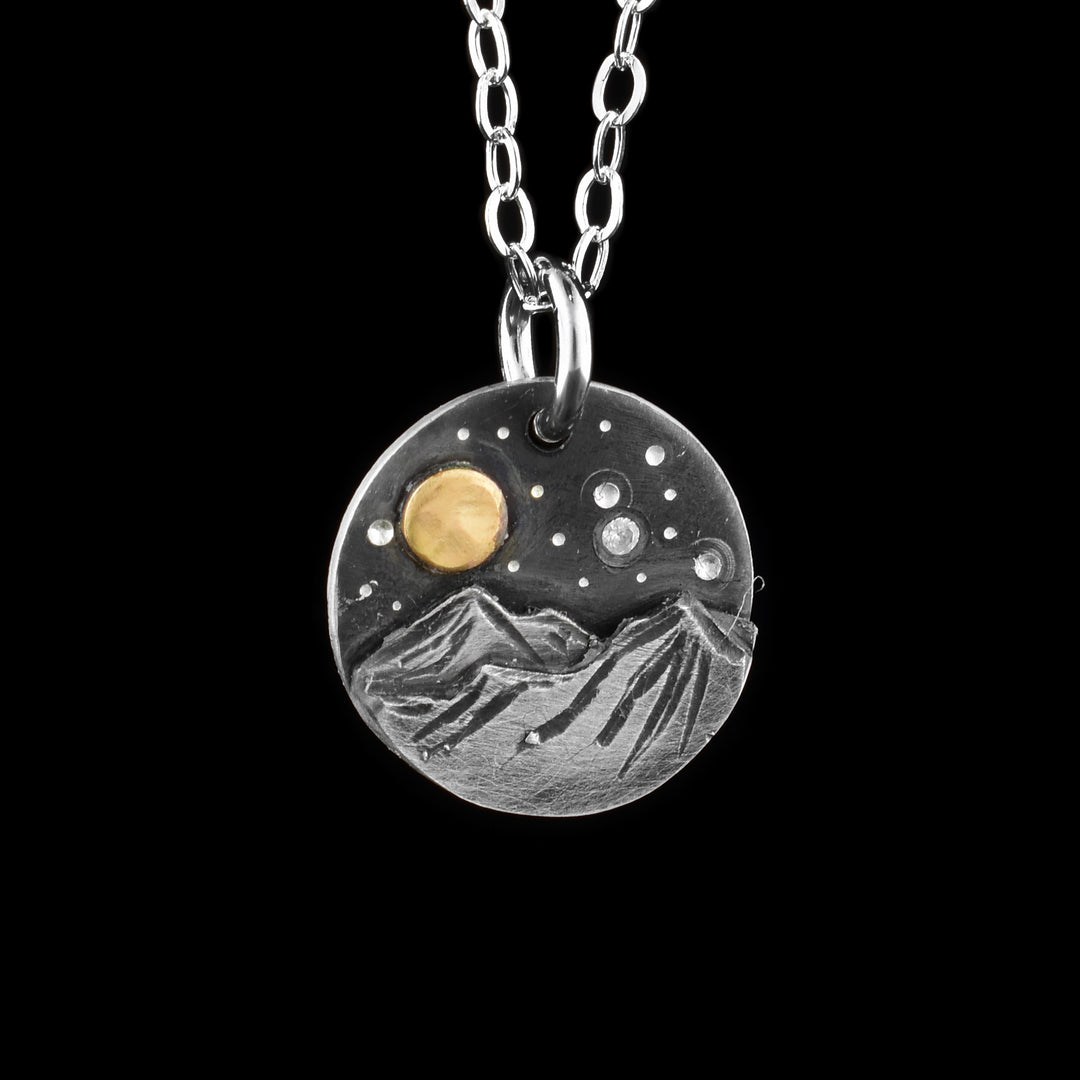 This mountain pendant necklace has a small, circle pendant dangling from a jump ring on a simple chain. These piece are made from sterling silver and there are mountains with a night sky above them on the pendant. There are two mountain peaks at the bottom that are textured and have 3D elements. Above the mountains there is a gold filled circle (moon) with silver dimples representing stars. There is one star that is a cubic zirconia gem.