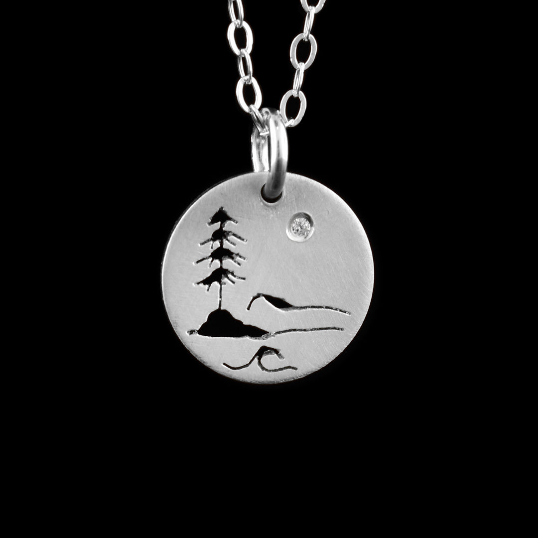 This Ocean pendant necklace has a small, circle pendant dangling from a jump ring on a simple chain. These pieces are made from sterling silver and there is a silhouette of Stanley park in Vancouver on the pendant. There is a tree on a rock with wave designs made around the rock and tree. At the top right, there is a cubic zirconia gem in the "Sky".