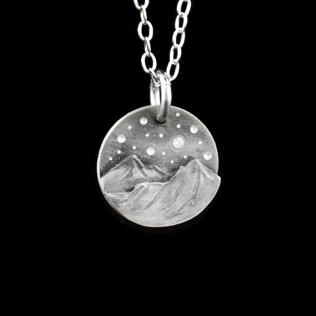 This mountain pendant necklace has a small, circle pendant dangling from a jump ring on a simple chain. These piece are made from sterling silver and there are mountains with a night sky above them on the pendant. There are two mountain peaks at the bottom that are textured and have 3D elements. Above the mountains, there are silver dimples representing stars and one star that is a cubic zirconia gem.