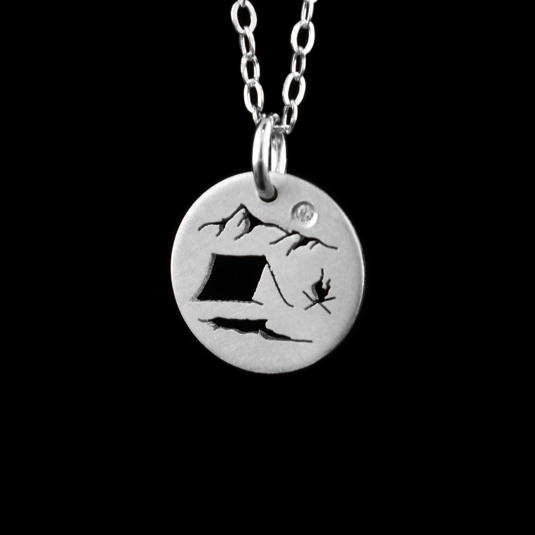 This camping mountain pendant necklace has a small, circle pendant dangling from a jump ring on a simple chain. These pieces are made from sterling silve and there is a silhouette scene of a campsite. There is a simple tent at the bottom with a campfire to the right of it. At the top of the piece, there are two mountain peaks with a cubic zirconia gemstone in the "Sky".