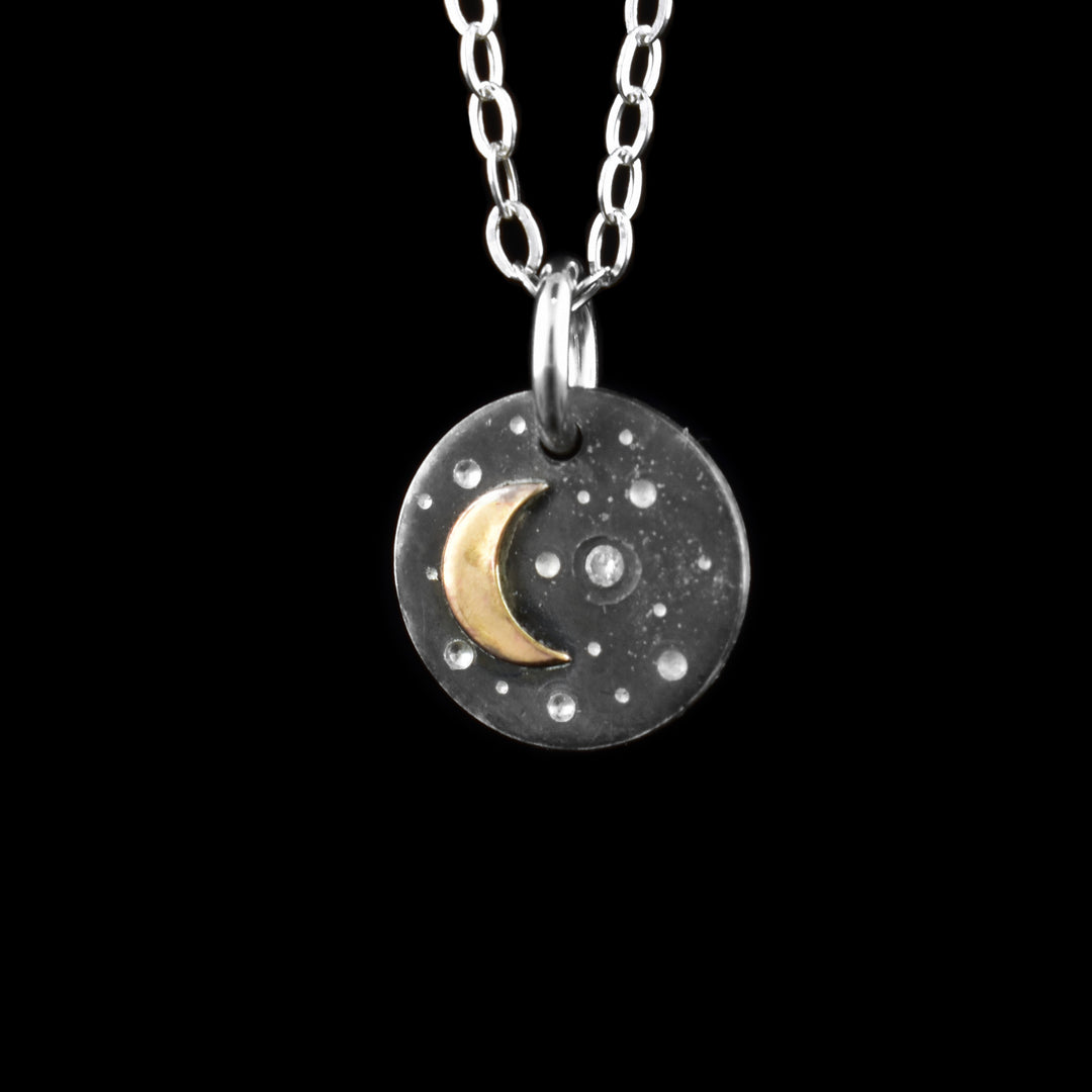 This gold crescent moon pendant necklace has a small, circle pendant dangling from a jump ring on a simple chain. These pieces are made from sterling silver and the pendant shows a crescent moon in a starry night sky. The moon has been done using 14K gold filled material and there are dimples made around the pendant representing stars. One of the stars is a cubic zirconia gem.