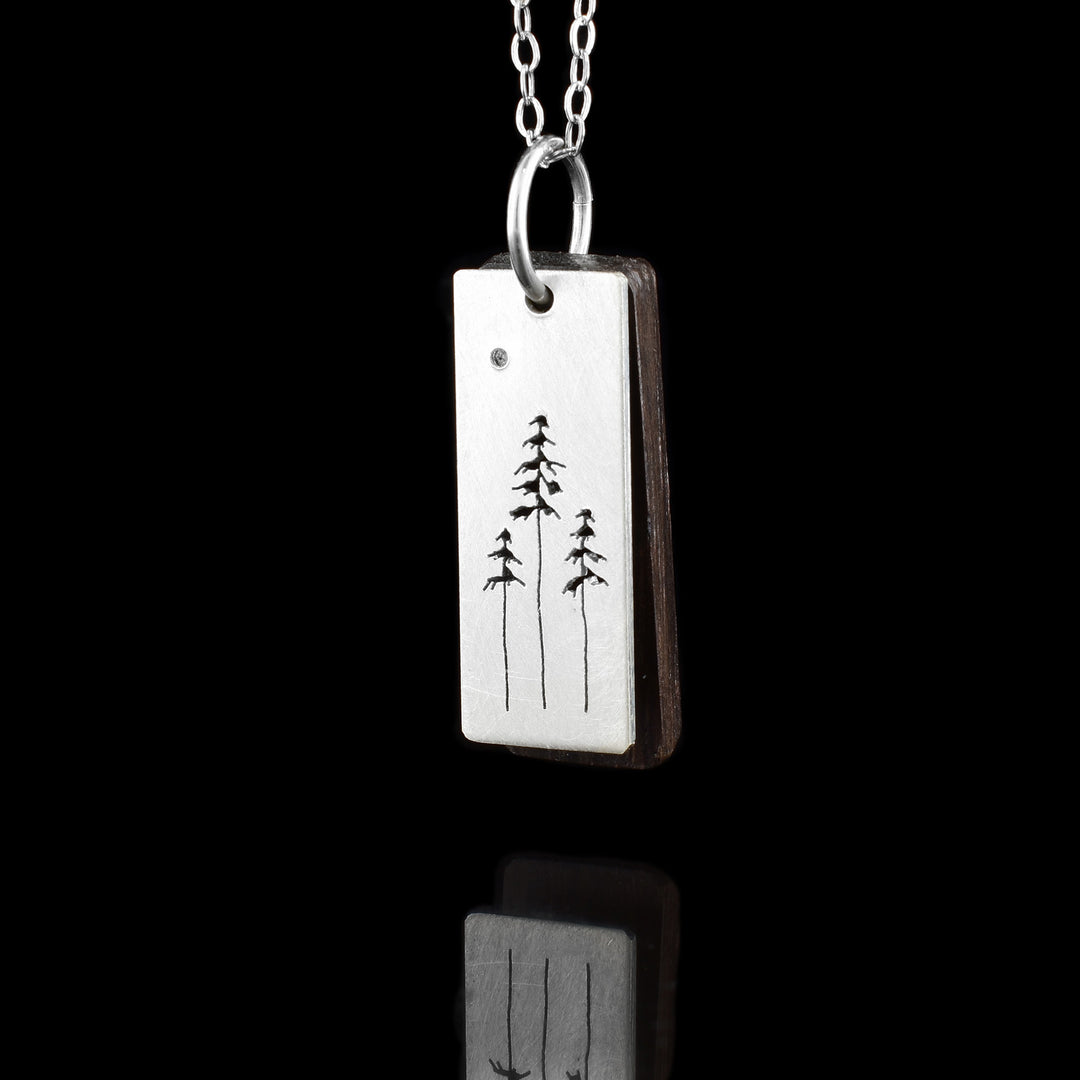 This hiker mountain pendant has a sterling silver rectangle pendant with a cutout pattern on it. There are three cedar trees with long trunks and patches of leaves closer to the tops of them. There is a dark, brown wooden piece behind the pendant that matches it in size. 