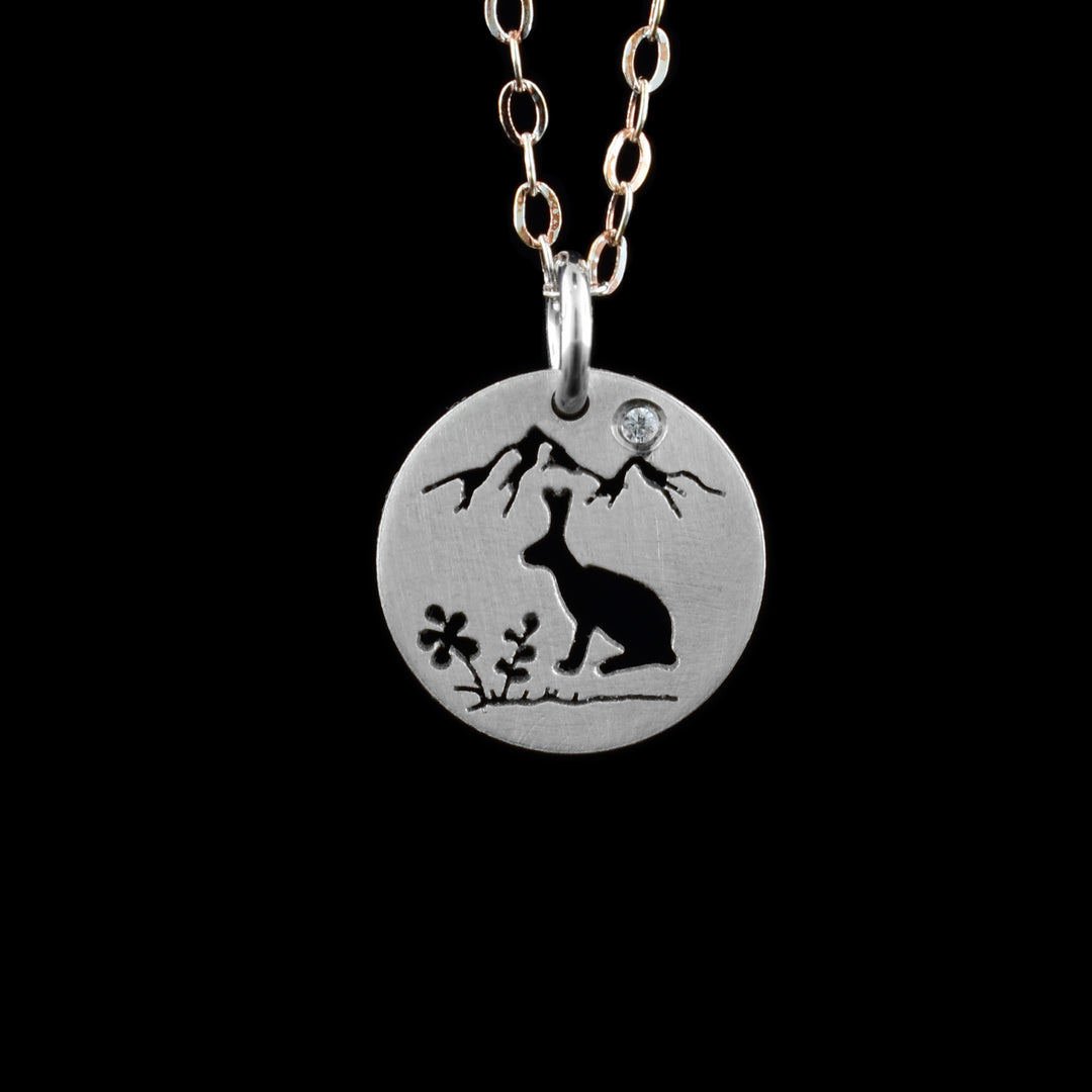 This bunny pendant necklace has a small, circle pendant dangling from a jump ring on a simple chain. These pieces are made from sterling silver and there is a silhouette scene of a bunny in a natural environment. The rabbit is shown to the right of the piece at a side profile and it is facing a small clump of wild flowers to its left. At the top of the pendant there are two mountain peaks with a cubic zirconia gem set in the "Sky".