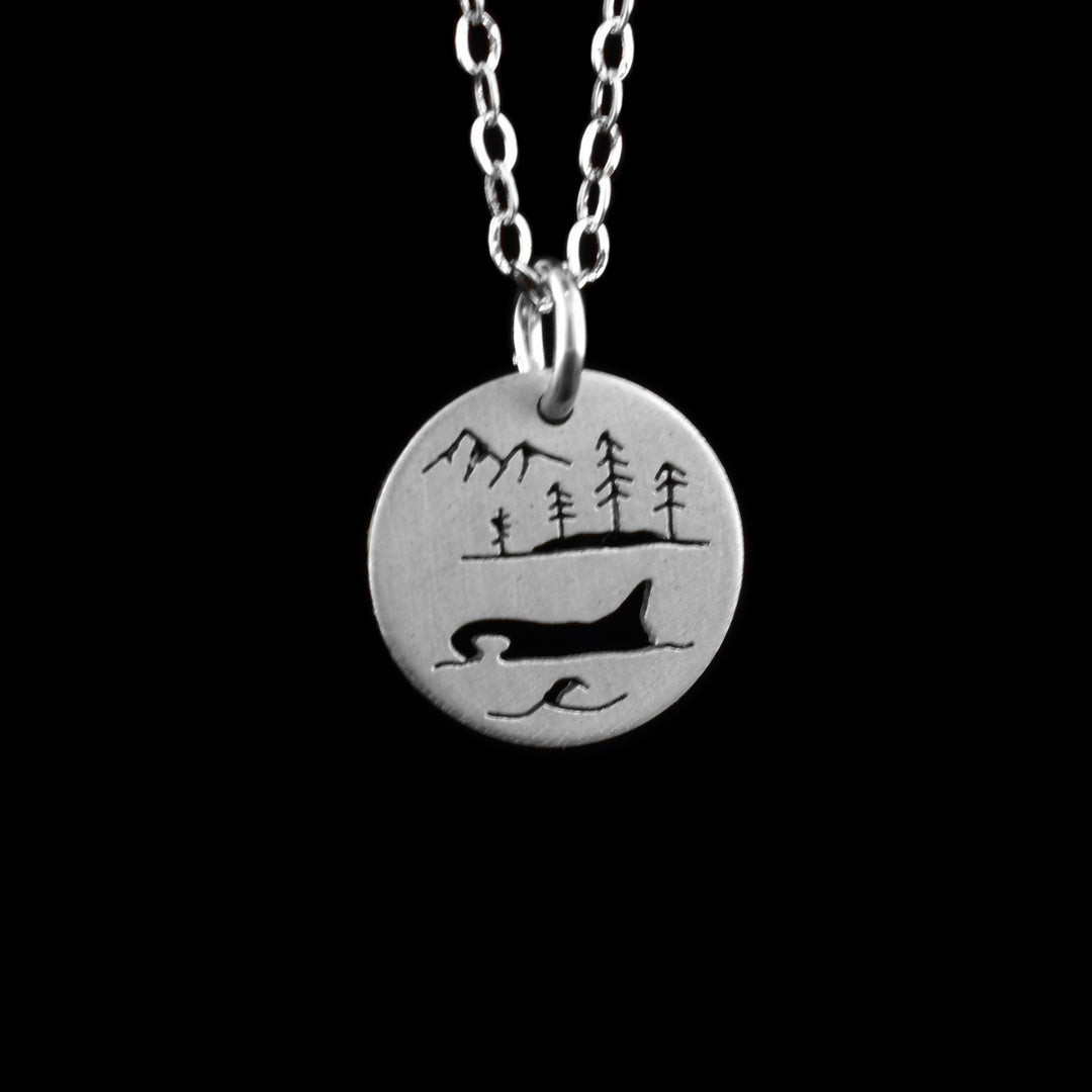This orca pendant necklace has a small, circle pendant dangling from a jump ring on a simple chain. These pieces are made from sterling silver and there is depiction of the orca in the west coast ocean. Half of the orca is visible through the water and there is an island with four simple trees on it above the whale. At the top of the piece are two mountain peaks.