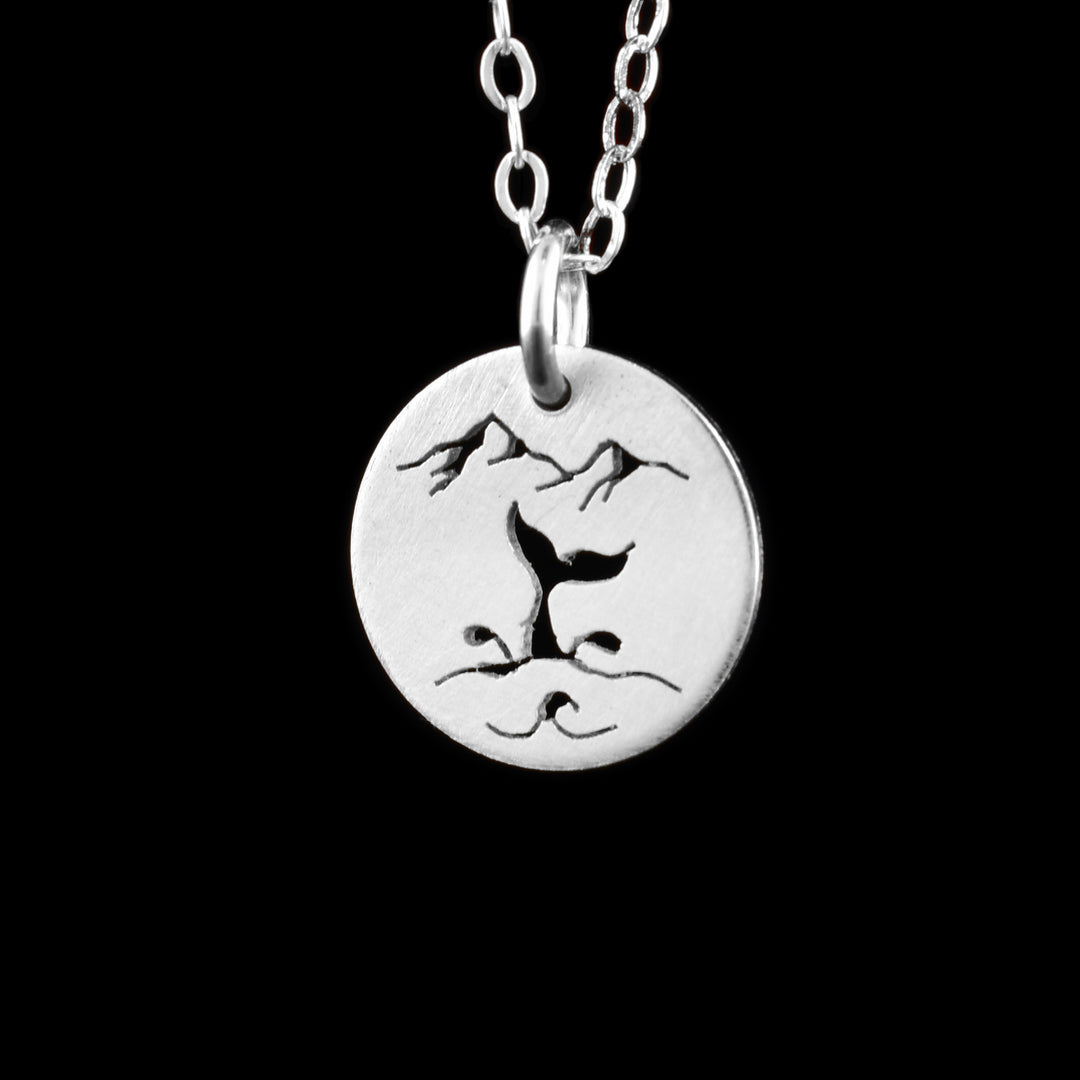 This whale tail pendant necklace has a small, circle pendant dangling from a jump ring on a simple chain. These pieces are made from sterling silver and there is a ocean scene on the pendant. There are silhouettes are two mountain peaks on top of the piece and there is a whale tail coming out from the waves with splashing designs coming out of the side.