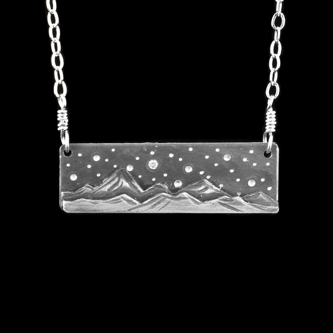 This mountain pendant necklace has a long, thin rectangle pendant that is horizontal. The chain is attached to both ends of the pendant which is made from sterling silver. There are textured and 3D mountain range at the bottom of the pendant. Above the mountains are dimples representing stars and a cubic zirconia gem set in the "Sky".