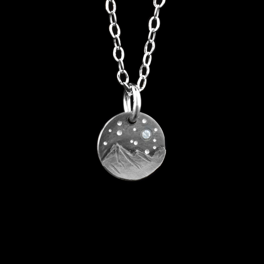 This mountain pendant necklace has a small, circle pendant dangling from a jump ring on a simple chain. These piece are made from sterling silver and there are mountains with a night sky above them on the pendant. There are two mountain peaks at the bottom that are textured and have 3D elements. Above the mountains, there are silver dimples representing stars and one star that is a cubic zirconia gem.