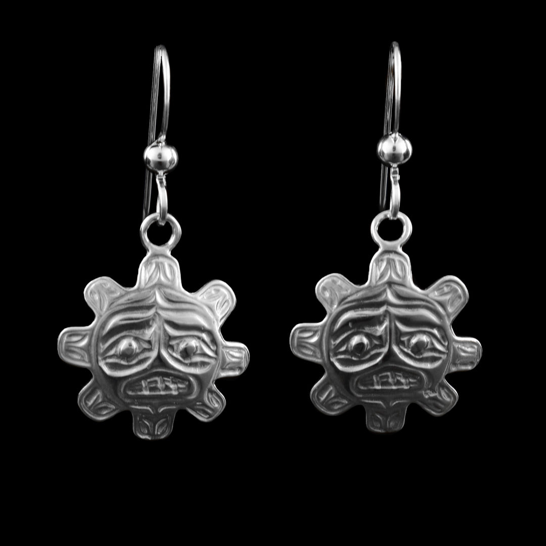 The Dangle earrings with fishhook ear wires, features a stylized sun face, expertly carved in sterling silver. The round face is encircled by radiating “rays”, giving it the classic sunburst shape. The face includes bold, expressive features arched brows, deep-set eyes, a prominent nose, and a defined mouth with visible teeth.