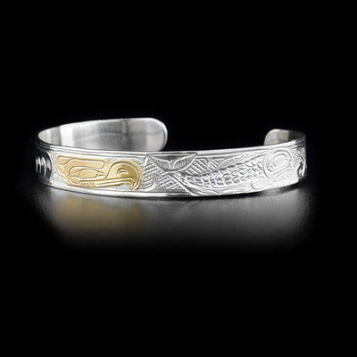 This eagle bracelet depicts a gold eagle head in the center facing a full-bodied salmon to its right. Waves have been carved in front of the salmon. Background has been cross-hatched.