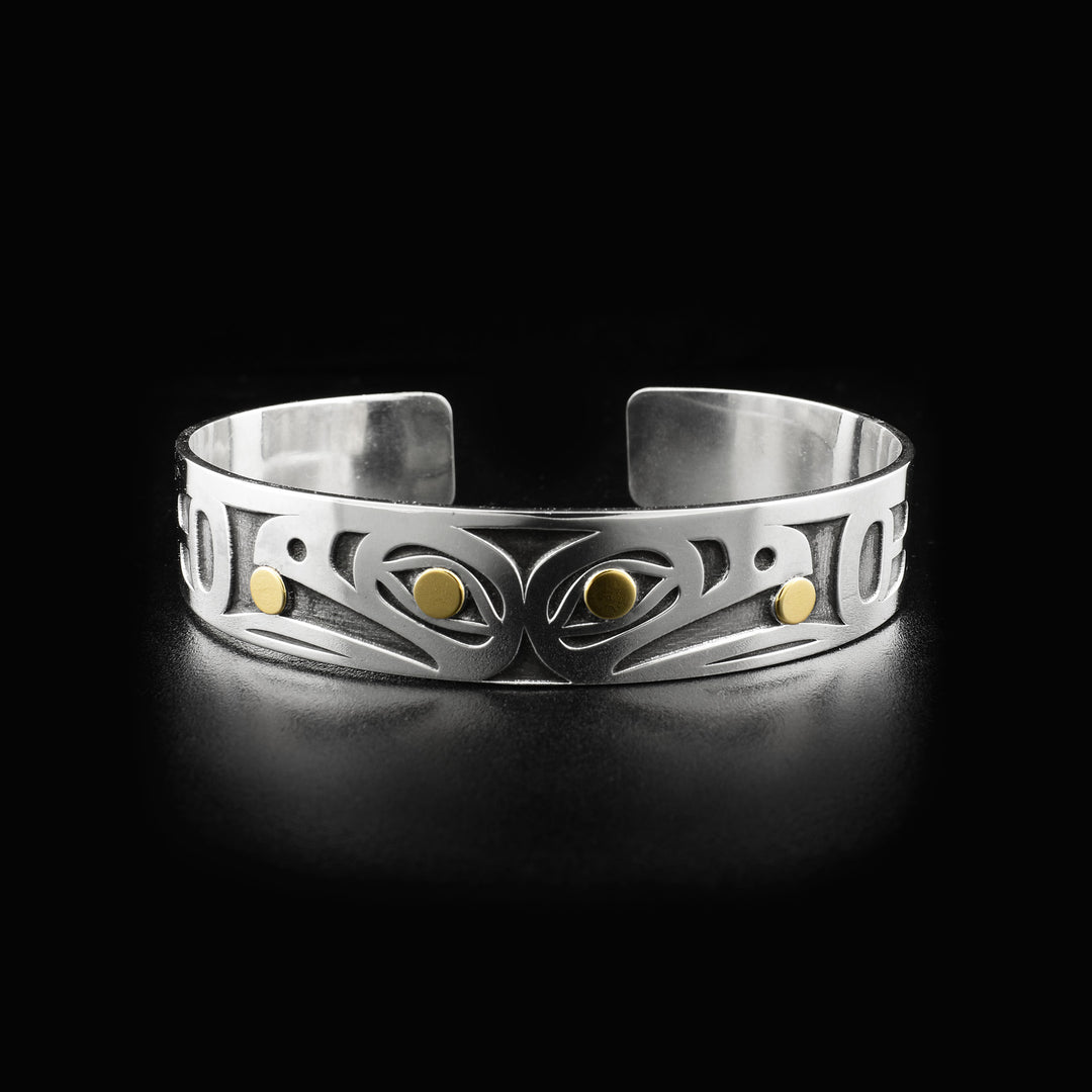 Side-view of two raven heads, back-to-back. Laser-carved with oxidized silver background. Both ravens have a 14K gold ball in their beak and 14K gold in their visible eye. Minimalist design, by Tahltan artist Grant Pauls. Bracelet is sterling silver.
