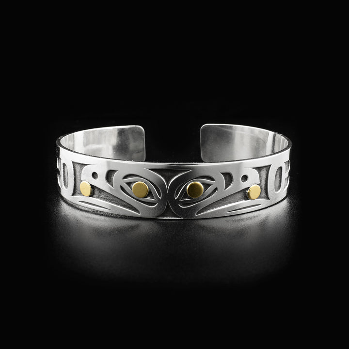 Side-view of two raven heads, back-to-back. Laser-carved with oxidized silver background. Both ravens have a 14K gold ball in their beak and 14K gold in their visible eye. Minimalist design, by Tahltan artist Grant Pauls. Bracelet is sterling silver.