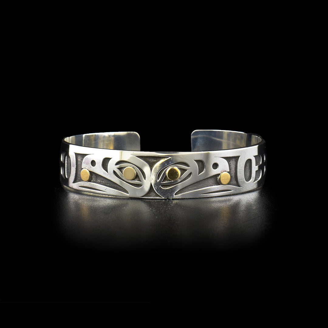 This photo of the silver and gold bracelet shows a side-view of two Raven heads back-to-back and facing outward. Laser-carved with oxidized silver background. Both ravens have a 14K gold ball in their beak and in their eye.