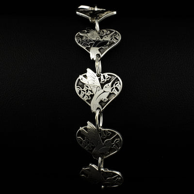 Heart-shaped frames with hummingbird flying among flowers inside. Design carved with laser. The oxidized background emphasizes the three-dimensional design. Material is sterling silver.
