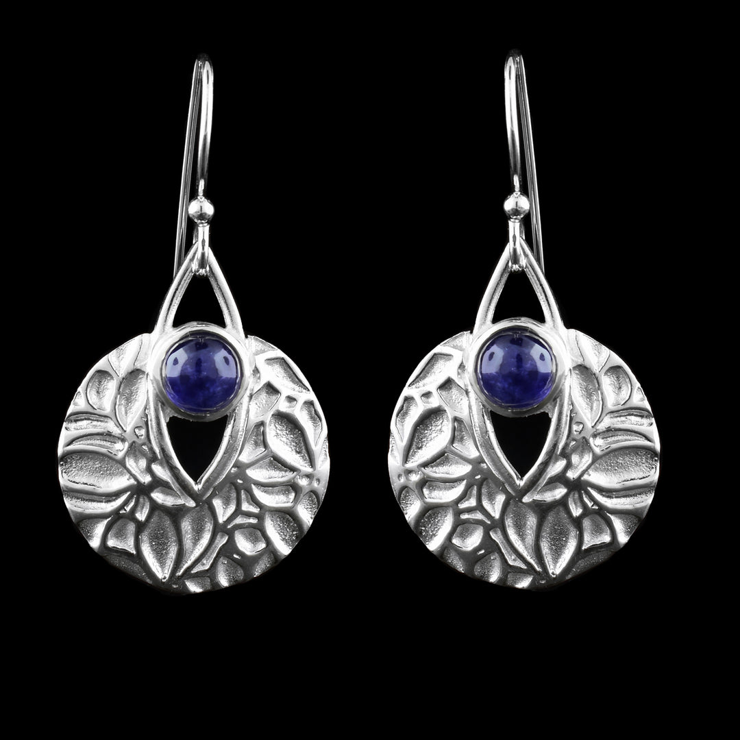 These handcrafted sterling silver gem dangle earrings feature a round, textured disc with an elegant floral and leaf pattern. At the center of each earring is a deep violet-blue iolite cabochon, adding a rich pop of color. The gemstone is framed by a graceful open circle design. The earrings hang from sleek French hook ear wires.