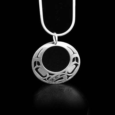 Round sterling silver cut-out pendant with pierced raven design. Raven’s head on bottom, facing left, with ball in beak. Designs on either side of the head depict wings. By Tahltan artist Grant Pauls.