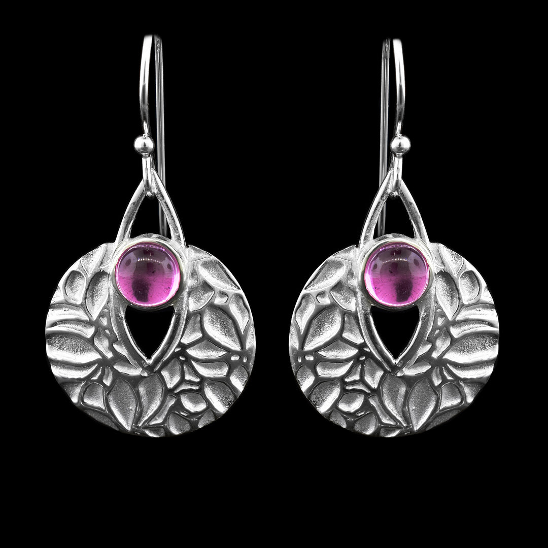 These handcrafted sterling silver gem dangle earrings feature a round, textured disc with an elegant floral pattern. At the top center of each earring is a pink sapphire. The gemstone is framed by a graceful open circle design. The earrings hang from sleek French hook ear wires.